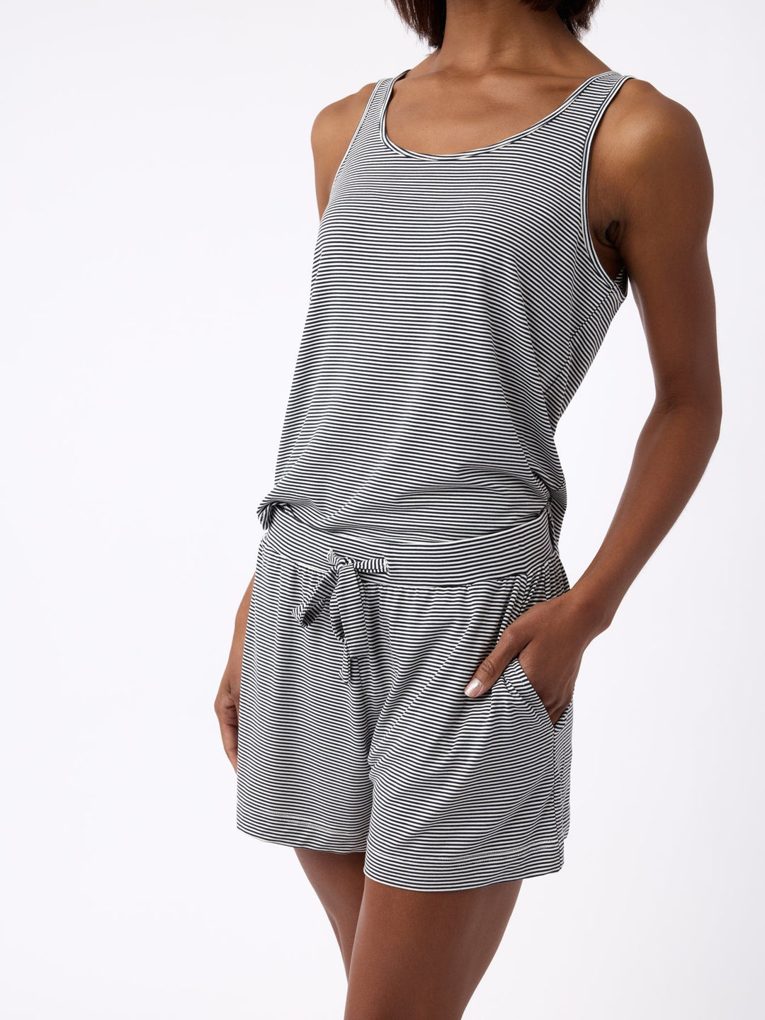 A person in Cozy Earth's Women's Bamboo Stretch-Knit Lounge Tank, a sleeveless gray and white striped romper with pockets, stands against a plain white background. The soft, comfortable fabric complements the casual pose with one hand in a pocket. |Color:Eclipse Mini Stripe